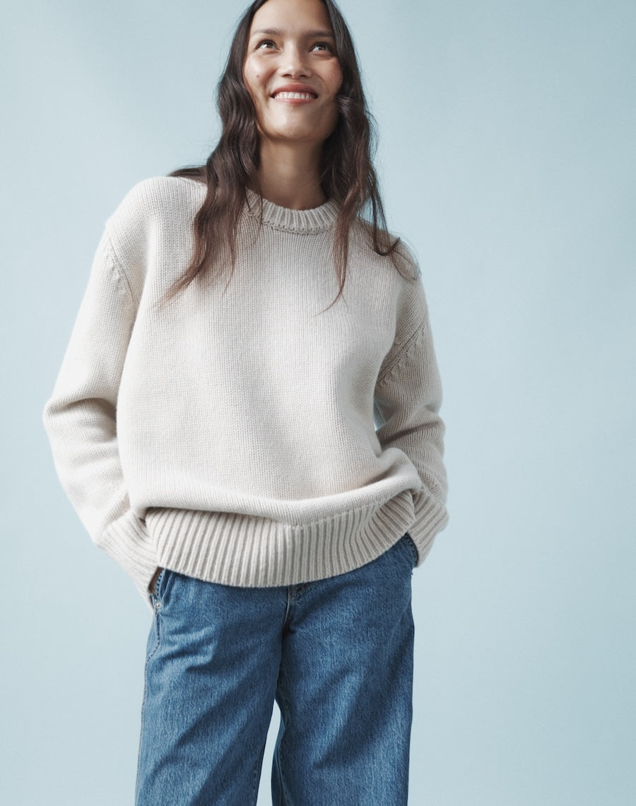 A woman wearing a beige oversized knit sweater with ribbed hem and neckline, paired with blue jeans.