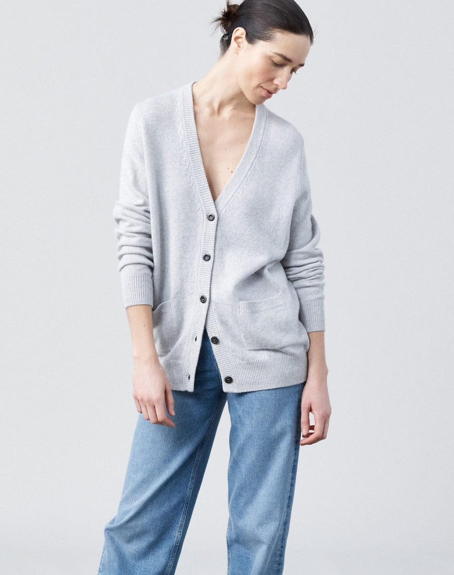 Woman wearing a light gray cardigan over a white top with blue jeans on a plain background