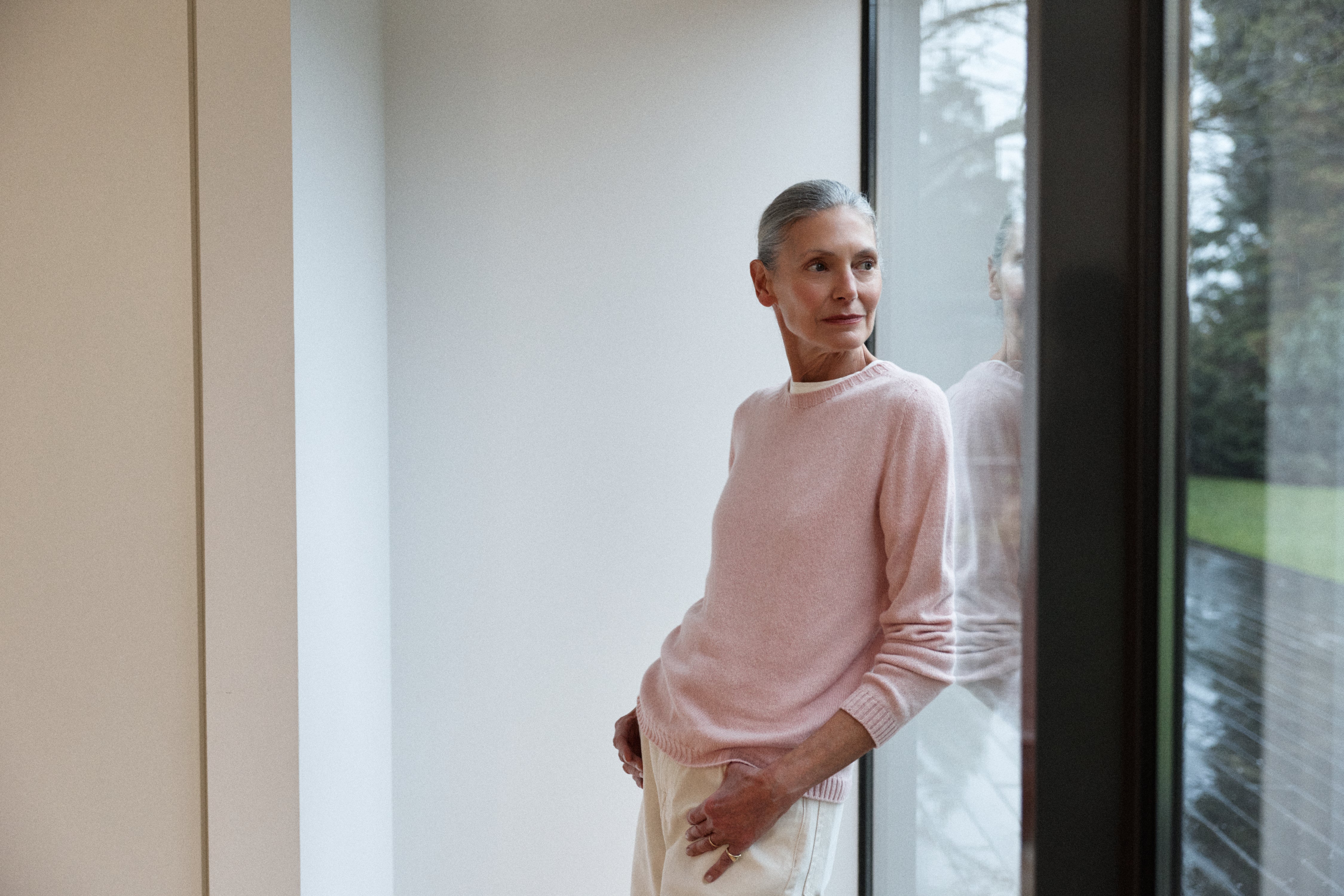 Woman in a pink sweater standing by a window with a view of trees.